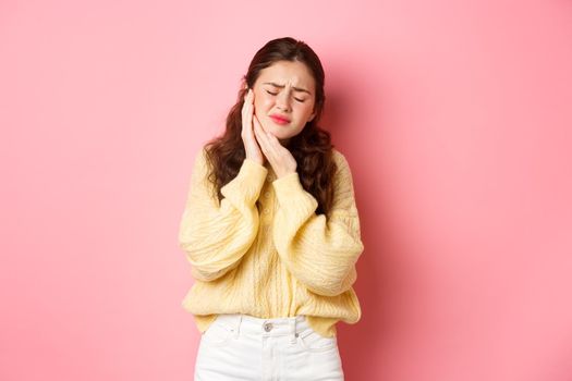 Young woman with terrible toothache, touching swallen cheek and grimacing from painful feeling, having tooth decay, need appointment to dentist, standing against pink background.