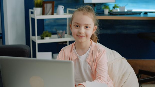 Portrait of schoolgirl using laptop for homework and smiling. Little child with modern gagdet for online remote education and courses looking at camera and feeling cheerful. Homeschooling