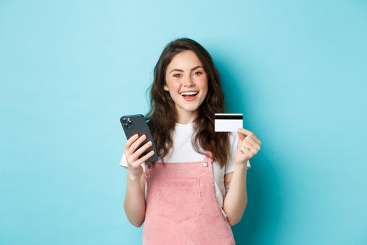 Stylish young woman online shopping, showing plastic credit card and holding smartphone, standing against blue background.