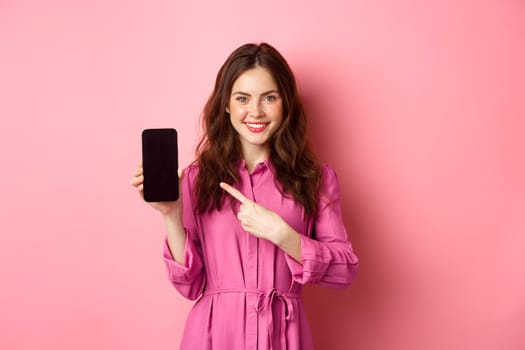 Young caucasian woman in stylish dress, pointing finger at smartphone screen and smiling, showing promo deal online, pink background.