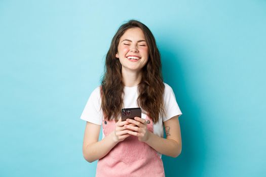 Portrait of happy young woman laughing with smartphone in hands, smiling and looking carefree, using mobile app, standing over blue background.