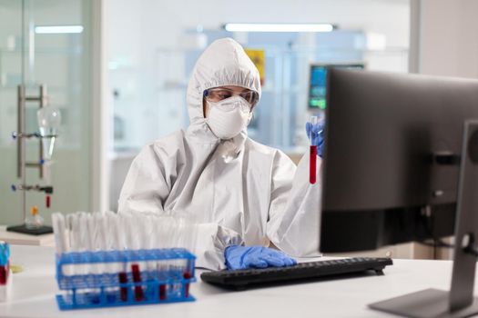 Medical researcher dressed in ppe suit looking at blood sample using computer. Scientist examining vaccine evolution using high tech technology for treatment development against covid19 virus.