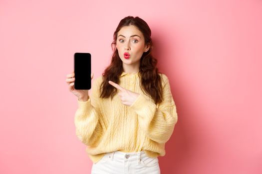 Technology and online shopping. Excited girl looks curious, points finger at empty mobile phone screen, shows app on smartphone, standing against pink background.