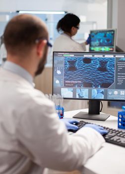 Man biologist checking DNA informations typing on pc in modern equipped laboratory. Group of biochemistry doctors examining virus evolution using high tech for researching treatment.