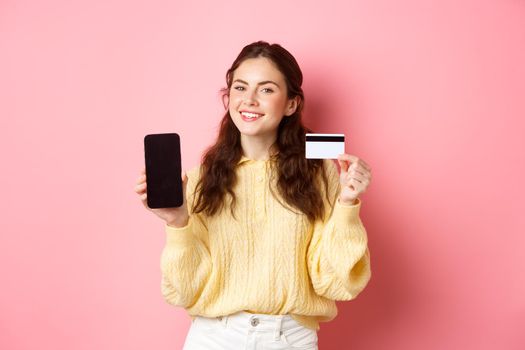 Technology and online shopping. Portrait of beautiful lady showing mobile phone screen and plastic credit card, smiling pleased, recommend app, standing over pink background.