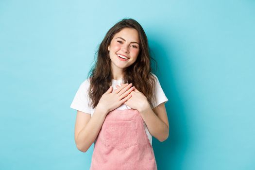 Portrait of grateful smiling young woman with curly hairstyle, summer make up, holding hands on heart and looking heartfelt, express gratitude, thanking you, blue background.