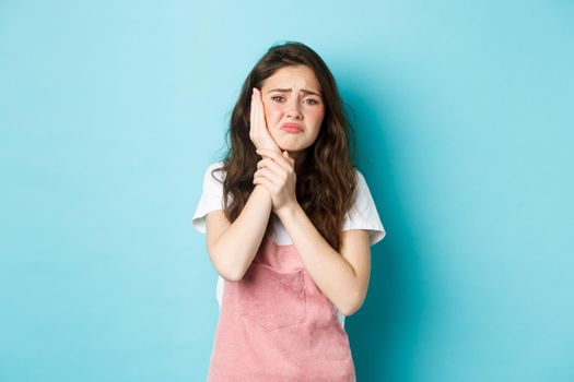 Image of frowning upset girl touching swallen cheek, having toothache, complaining on tooth decay, need to see dentist, standing against blue background.