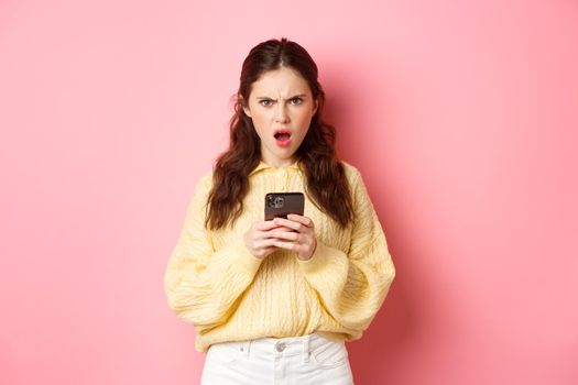 Confused and angry girlfriend reading strange message on phone, staring at camera frustrated and questioned, standing against pink background.