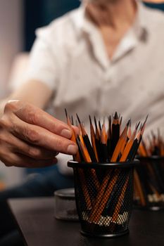 Close up of caucasian hand on colorful pencils for fine art and drawing on table in artwork studio room. Old person touching art intruments on desk at home. Artist with professional tools