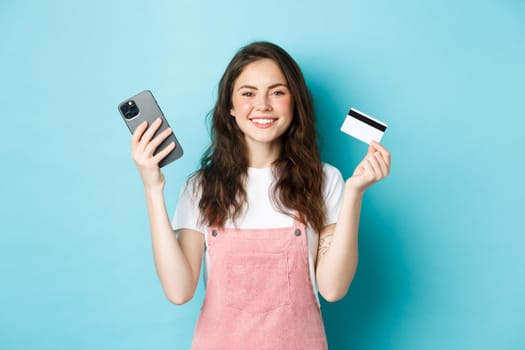 Smiling cute girl showing plastic credit card and smartphone, order something in store via mobile app, shop online, or pay for order, standing against blue background.