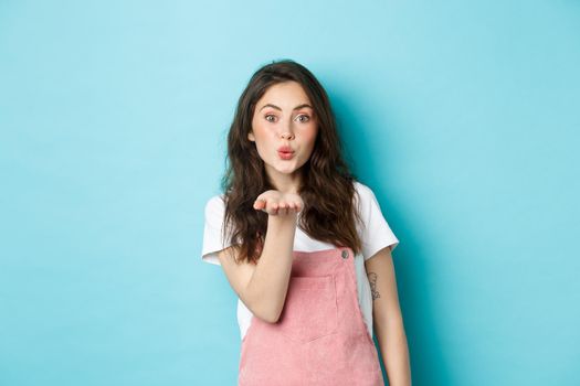 Beautiful girl blowing air kiss with romantic face, holding palm near lips and sending mwah at you, standing in summer outfit against blue background.
