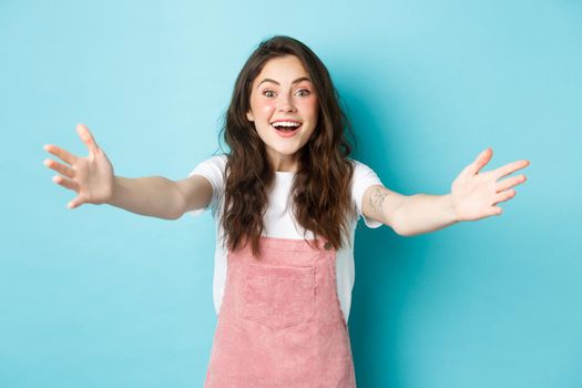 Image of happy young woman with curly hair and cute summer blush make up, stretch out hands to hug or hold someone, reaching for embraces, welcome you, blue background.