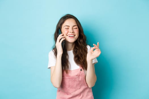 Portrait of cute and stylish girl laughing while chatting on phone, holding smartphone near ear and smiling happy, having casual conversation, standing over blue background.