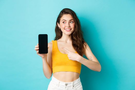 Portrait of cute brunette woman pointing finger at smartphone screen, looking left at copy space, standing in summer outfit against blue background.