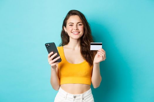Online shopping. Beautiful woman getting ready for summer vacation, booking tickets with credit card and smartphone app, standing over blue background.