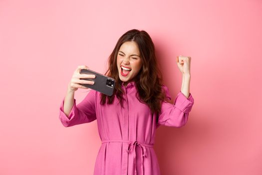 Technology concept. Excited girl winning on mobile phone video game, shouts yes and makes fist pump with satisfied expression, pink background.