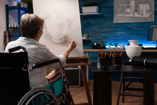 Old woman with disability creating drawing of vase from table in workshop space. Senior artist sitting in wheelchair while working on modern masterpiece in artwork studio