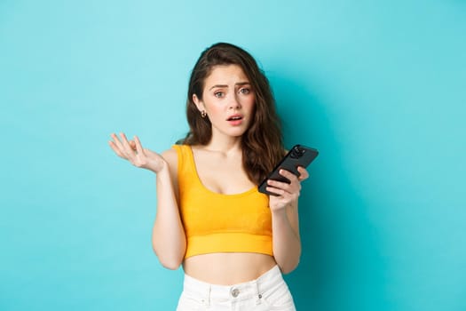 Technology and lifestyle concept. Glamour girl looks confused after reading smartphone screen, staring puzzled at camera, standing against blue background.