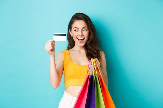 Image of happy woman shopaholic showing her plastic credit card, holding shopping bags, wearing summer clothes, standing against blue background.