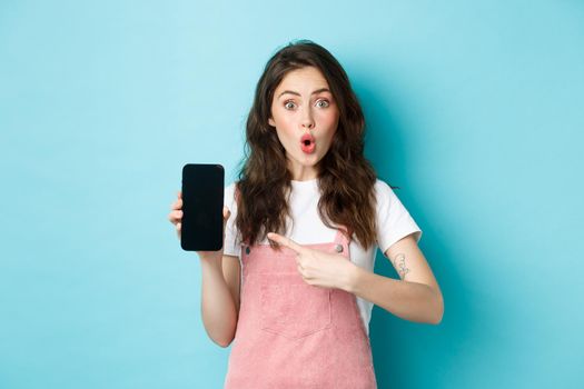 Impressed young modern woman pointing at smartphone screen, say wow and stare amazed at camera, showing awesome application, new online store, standing over blue background.