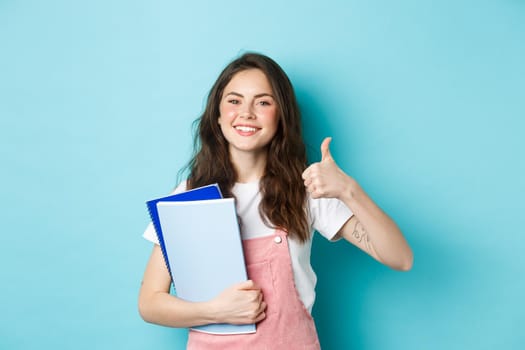 Young woman attend courses, girl student studying, holding notebooks and showing thumb up in approval, recommending company, standing over blue background.