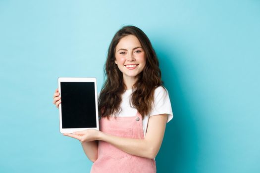 Portrait of smiling beautiful woman demonstrate blank tablet screen, showing promo on device screen, standing against blue background.