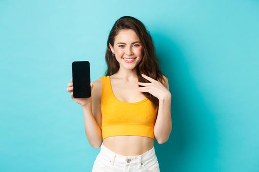 Stylish caucasian woman in summer outfit showing empty smartphone screen and smiling, demonstrate an app or online store, standing over blue background.
