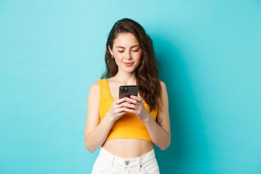 Stylish young woman checking messages on phone, reading smartphone screen and smiling, shopping online, standing over blue background.