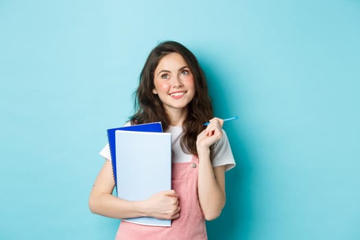 Beautiful girl student looking thoughtful at top copy space, standing with notebooks homework and smiling pensive, standing against blue background.