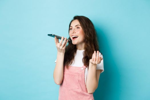 Young woman using app translator on smartphone, speaking into mobile phone dynamic, holding cellphone near lips, recording voice message, blue background.