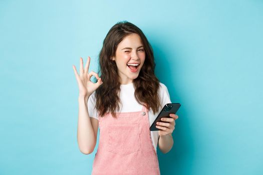 Online shopping. Cheerful cute girl winking at you, smiling and showing okay sign after using smartphone app, recommending internet shop or social media page, blue background.