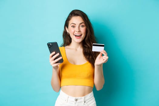 Young happy woman making online order, shopping with credit card and smartphone, look amazed at camera, standing over blue background.