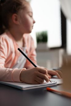 Closeup of little child writing mathematics homework on notebook during classroom lesson sitting at desk table in living room. Concept of home schooling, distance learning, online education