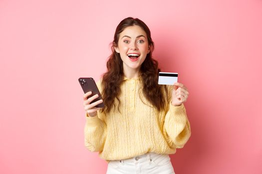 Technology and online shopping. Excited girl making order, paying online with plastic credit card, holding mobile phone and smiling at camera, pink background.