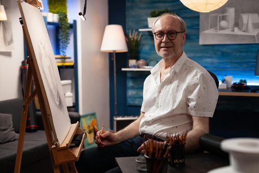 Portrait of caucasian elder artist sitting in artwork studio. Senior man with artistic imagination looking at camera while working on vase drawing. Aged person with masterpiece on canvas