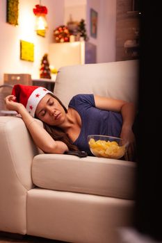 Woman with santa hat sleeping on couch after watching winter entertainment movie on television in xmas decorated kitchen. Caucasian female celebrating december season enjoying christmas holiday