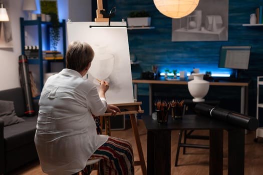 Elder woman drawing vase design from table on canvas while sitting in artwork studio. Senior artist using professional artistic tools to create and draw masterpiece in workspace room