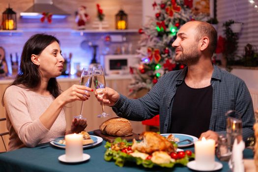 Happy family clinking glass of wine sitting at dining table celebrating christmas traditional holiday. Romantic cheerful couple spending christmastime together enjoying winter season