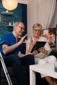 Elder married couple talking on video call conference for remote online communication using digital tablet while sitting on living room sofa. Old husband and wife with internet technology