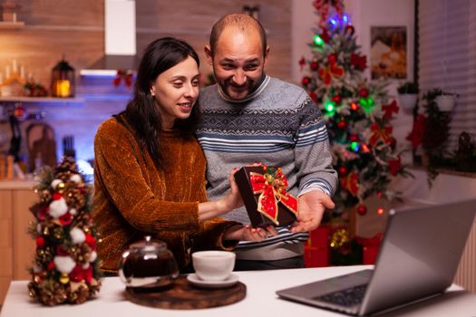 Happy family showing xmas present surprise to remote friends during online videocall meeting on laptop computer standing in festive decorated kitchen. Cheerful couple enjoying christmas holiday