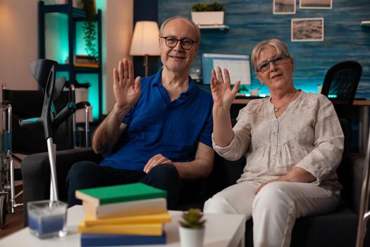 POV of elder couple waving hands on video call conference in living room. Senior people looking at camera while sitting on sofa and using online remote communication. Old husband and wife