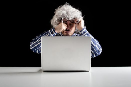 Portrait of a senior man rubbing his tired eyes sitting in front of the laptop computer on a white table. Black background