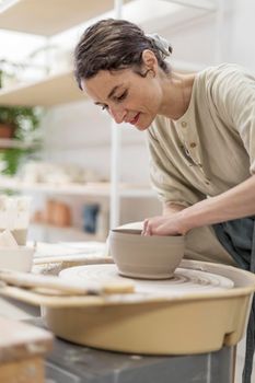 Young woman holding pottery instrument for scraping, smoothing, shaping and sculpting. Lady siting on bench with pottery wheel and making clay pot