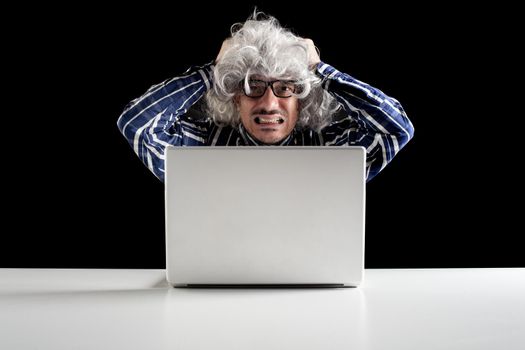 Portrait of a stressed senior man working on a laptop computer sitting at a white table on black background