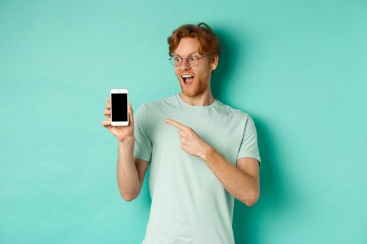 Check this out. Handsome redhead guy in glasses pointing finger at blank smartphone screen, showing online promotion, standing amazed over turquoise background.