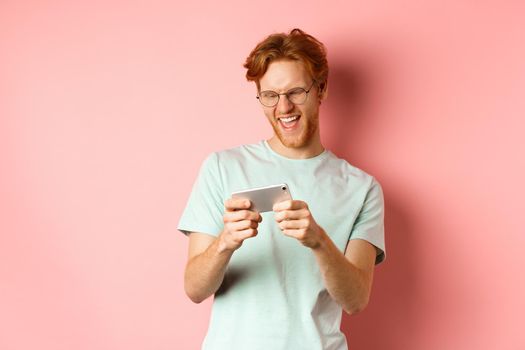 Happy young man with red messy haircut, wearing glasses, playing video game on smartphone and having fun, looking at mobile screen, standing over pink background.