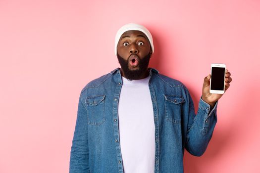 Amazed Black guy in streetstyle clothes showing mobile phone screen, saying wow as demonstrating smartphone display, standing over pink background.