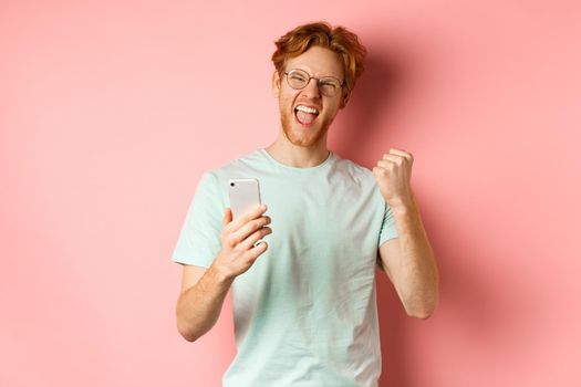 Happy redhead guy in glasses and t-shirt winning online prize, shouting yes with joy and satisfaction, holding smartphone and making fist pump, pink background.