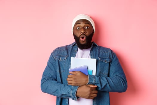 Education. Excited african-american adult student carry notebooks, staring at camera amaze, standing over pink background.