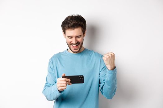 Happy young man winning in video game on smartphone, looking at mobile screen and say yes, making fist pump in celebration, achieve online goal, standing over white background.